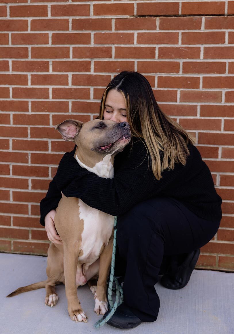 female staff member kneeling down to hug and kiss dog outside in front of red brick wall
